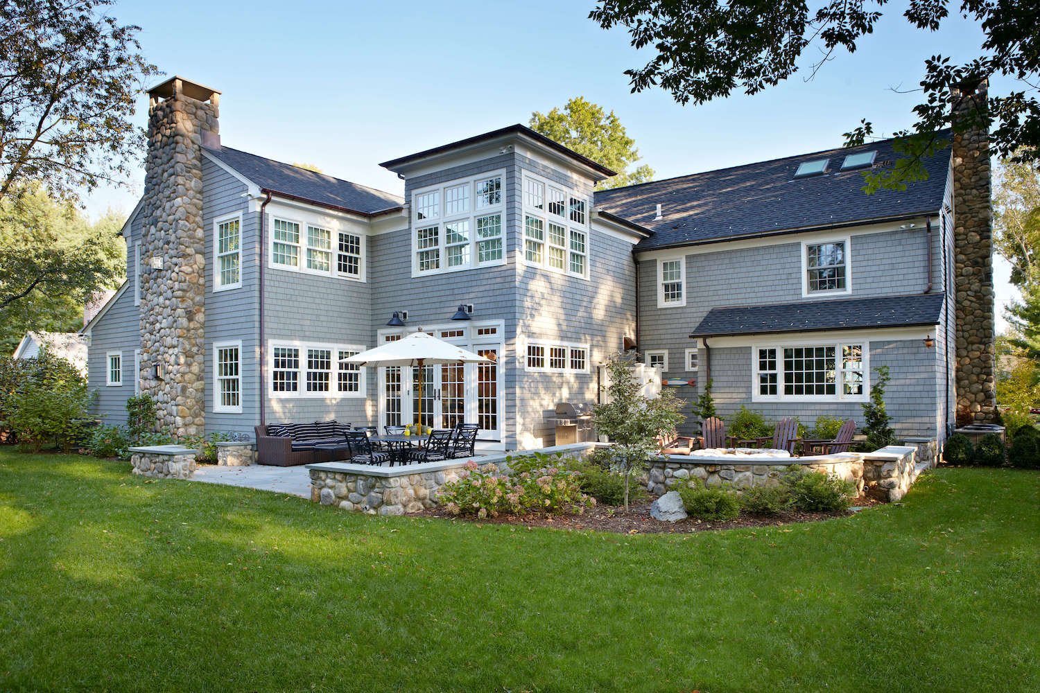A large custom home with blue siding, a stone chimney, and french doors opening onto a patio surrounded by a stone wall by TR Design-Build