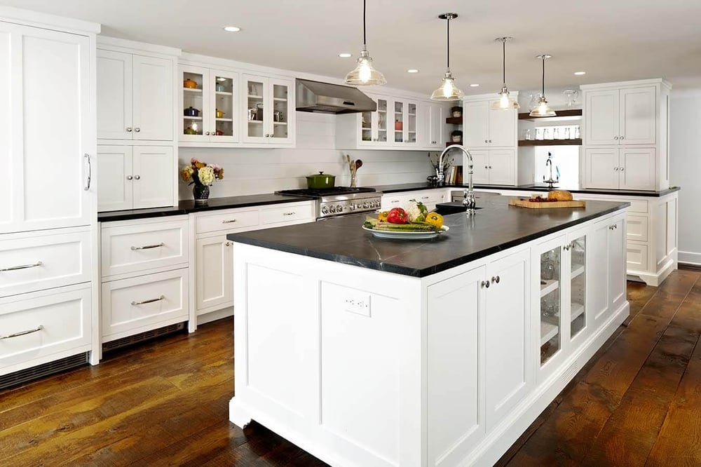 A remodeled kitchen with a large island with a sink, stone countertops, hardwood floors, and white cabinetry by TR Design-Build