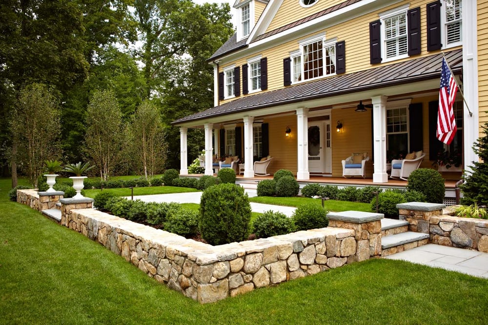 Front view of a New Canaan exterior home remodel with yellow siding and black shutters, featuring a covered porch and a patio surrounded by a stone wall.